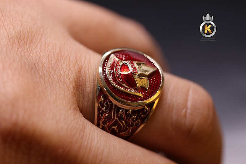 May include: A gold ring with a red enamel design featuring a stylized warrior helmet. The ring has intricate details and a polished finish.