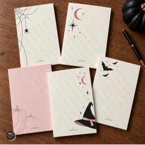 Peut inclure: Cinq blocs-notes rectangulaires en papier présentant divers motifs, notamment une toile d'araignée avec une araignée, un croissant de lune avec des étoiles, des citrouilles et un chapeau de sorcière. Chaque bloc est crème ou rose clair avec le texte JADGS.