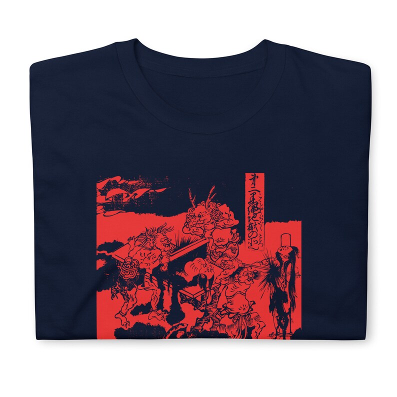 May include: A navy blue t-shirt with a red print of a Japanese woodblock print depicting a scene with multiple figures. The print is in a traditional style and features a red background with black outlines. The text on the right side of the print reads "The Tale of the Great Fire of the Capital".