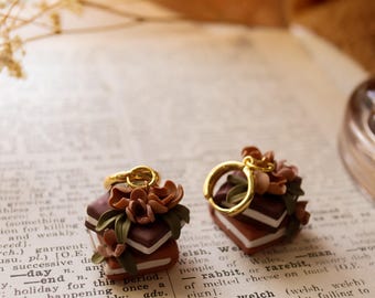 May include: A pair of unique earrings featuring a miniature stack of books with brown and white pages, topped with brown flowers and a gold hoop. The earrings are set against a vintage book page.