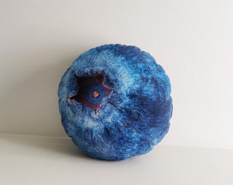 May include: A round decorative pillow shaped like a blueberry. The pillow features a detailed print of a blueberry's skin, with shades of blue and a red star-shaped center. The pillow is placed on a white surface.