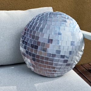 Könnte beinhalten: Ein runder, silberner Kissenbezug im Disco-Kugel-Design mit Mosaikfliesen. Das Kissen liegt auf einem hellgrauen Kissen, im Hintergrund ein weiteres graues Kissen. Das Kissen ist ein dekorativer Artikel.
