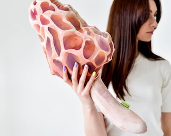 May include: A large, decorative mushroom-shaped pillow with a brown and orange patterned design. The pillow is being held by a person with their hand visible.