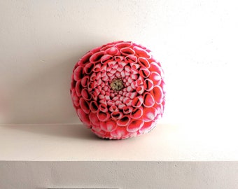 Op de afbeelding: Een rond roze en wit bloemenkussen met een dahlia-ontwerp.