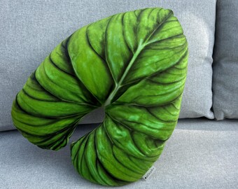 Op de afbeelding: Een decoratief kussen in de vorm van een groot groen blad met gedetailleerde nerven. Het kussen heeft een levendige groene kleur en ligt op een grijze ondergrond. Het blad-vormige kussen voegt een vleugje natuurlijke decoratie toe.