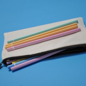 Peut inclure: Une trousse en toile de couleur cr&egrave;me avec une fermeture &eacute;clair noire, ouverte pour r&eacute;v&eacute;ler plusieurs crayons aux couleurs pastel. Les crayons sont dans des tons de bleu, jaune et rose. Le fond est bleu clair uni.