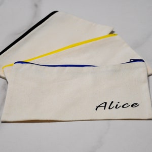 Peut inclure: Trois pochettes zipp&eacute;es en toile &eacute;crue avec garnitures color&eacute;es. L'une a une garniture de fermeture &eacute;clair noire, une autre jaune et la troisi&egrave;me bleue. Le mot "Alice" est imprim&eacute; en noir sur le devant d'une pochette.