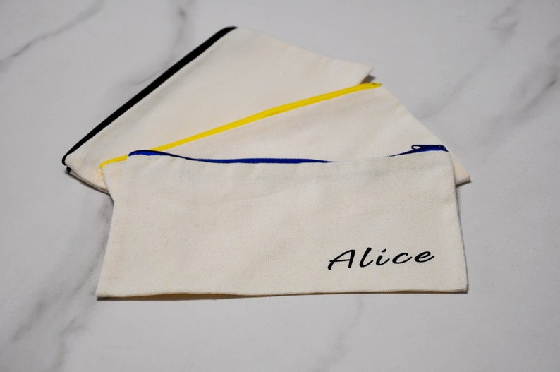 Peut inclure: Trois pochettes zipp&eacute;es en toile &eacute;crue avec garnitures color&eacute;es. L'une a une garniture de fermeture &eacute;clair noire, une autre jaune et la troisi&egrave;me bleue. Le mot "Alice" est imprim&eacute; en noir sur le devant d'une pochette.