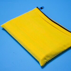 Peut inclure: Une pochette rectangulaire jaune vif avec une fermeture &eacute;clair et une bordure noires. La pochette est en tissu textur&eacute;, pos&eacute;e sur un fond bleu uni. La tirette de la fermeture &eacute;clair est argent&eacute;e.