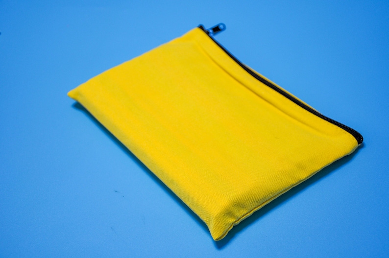 Peut inclure: Une pochette rectangulaire jaune vif avec une fermeture &eacute;clair et une bordure noires. La pochette est en tissu textur&eacute;, pos&eacute;e sur un fond bleu uni. La tirette de la fermeture &eacute;clair est argent&eacute;e.