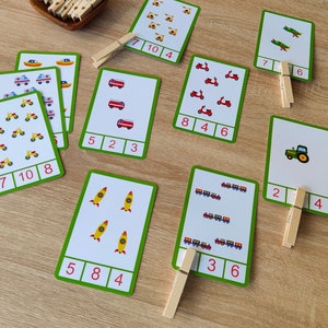 May include: A set of ten colorful flashcards with various images of vehicles, including airplanes, fire trucks, motorcycles, tractors, rockets, and trains. Each card has a number printed on it, and there are wooden clothespins to help with counting.