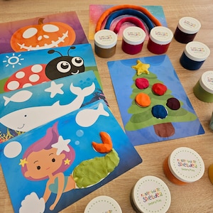 Könnte beinhalten: Ein Set mit bunten Knetmassen mit verschiedenen Themen, darunter ein Kürbis, ein Marienkäfer, ein Wal, eine Meerjungfrau, ein Weihnachtsbaum und ein Regenbogen. Die Knetmassen befinden sich in Behältern mit dem Text "K mie bu te Spielide" und "Sensory Play".