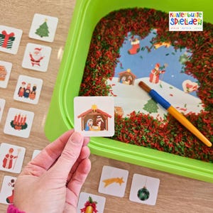 Op de afbeelding: Een hand houdt een vierkante kerstkaart vast met een afbeelding van de kerststal. Andere kaarten met kerstthema-illustraties liggen verspreid op een houten oppervlak. Een groene bak gevuld met rode en groene rijst en een geschilderde winterscène staan op de achtergrond.