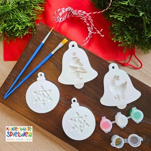 Puede incluir: Un conjunto de cuatro adornos navideños de madera blanca con diferentes diseños: una campana con un árbol de Navidad, una campana con un reno y dos adornos con copos de nieve. Los adornos están sobre una superficie de madera con dos pinceles y una bolsa de cuerda. También hay cuatro pequeños botes de pintura roja, blanca, dorada y verde.