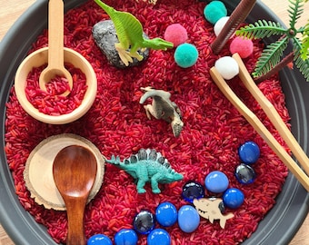 Puede incluir: Un contenedor sensorial rojo lleno de arroz rojo, gemas de vidrio azul y juguetes de dinosaurios de plástico. También hay pinzas de madera, una cuchara de madera y un pequeño tazón de madera con arroz rojo en él. El contenedor está sobre una superficie negra.