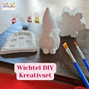 Puede incluir: Un kit de manualidades DIY que incluye un gnomo blanco, un copo de nieve, una puerta y dos pinceles. El kit está etiquetado como "Wichtel DIY Kreativset".