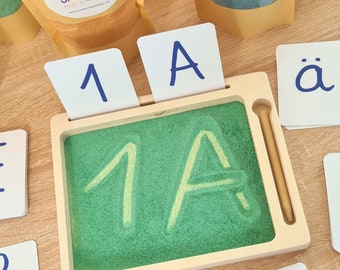 May include: A wooden tray with a green sand surface and a wooden stylus. The tray has a slot for cards, and there are cards with the numbers 1 and 3, the letters A and ä, and the number 1A written on them.