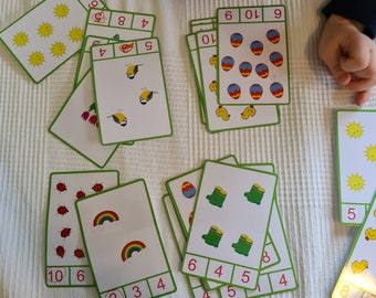 May include: A collection of educational playing cards with green borders, each featuring colorful illustrations of items like suns, birds, eggs, ladybugs, rainbows, and boots, alongside numerical sequences and red handwritten numbers in boxes.