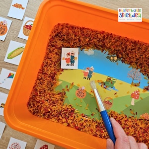 Op de afbeelding: Een oranje plastic bak gevuld met oranje gekleurde rijst en herfstthema-illustraties. De bak bevat een penseel met een blauwe handgreep en verschillende kaarten met herfstafbeeldingen. De tekst "Trick or Treat" is zichtbaar.