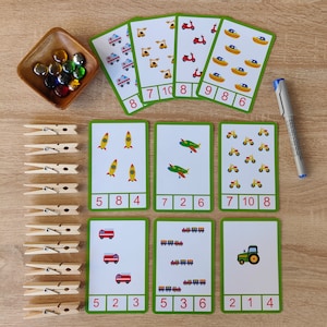 May include: A set of 10 green and white flashcards with colorful illustrations of rockets, airplanes, motorcycles, fire trucks, trains, and tractors. Each card has a number from 1 to 10 and a corresponding number of images. There are also 15 wooden clothespins.