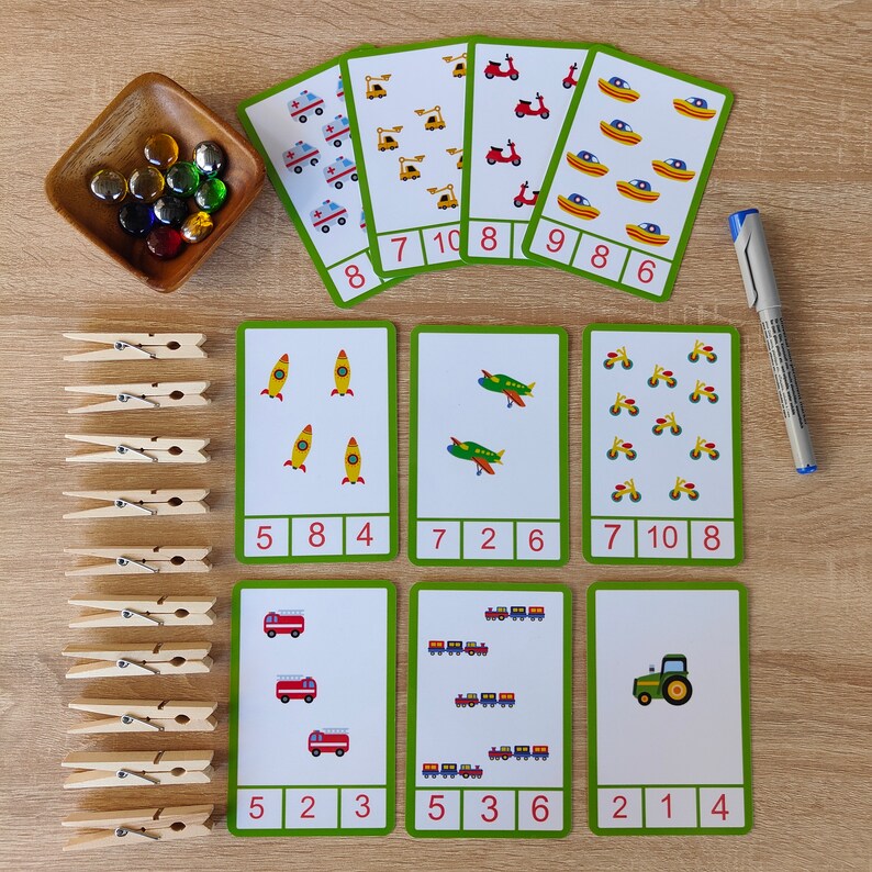 May include: A set of 10 green and white flashcards with colorful illustrations of rockets, airplanes, motorcycles, fire trucks, trains, and tractors. Each card has a number from 1 to 10 and a corresponding number of images. There are also 15 wooden clothespins.