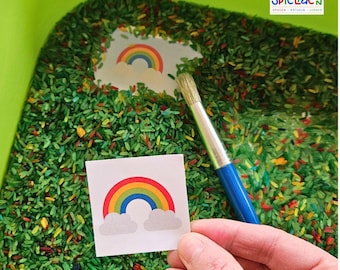 May include: A rainbow cutout on a white card is held above a green, yellow, and red rice sensory bin. A blue paintbrush is also in the bin. The text "Kunterbunte SpielIdeen" is visible in the top right corner of the image.