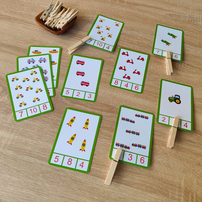 May include: A set of ten colorful flashcards with various images of vehicles, including airplanes, fire trucks, motorcycles, tractors, rockets, and trains. Each card has a number printed on it, and there are wooden clothespins to help with counting.