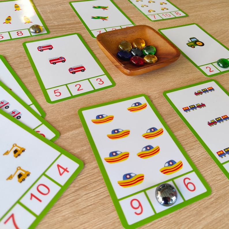 May include: A set of colorful counting cards featuring various images, including rockets, fire trucks, boats, tractors, and trains. Each card has a number from 1 to 10 printed on it, along with the corresponding number of images. The cards are laid out on a wooden surface with some glass gems scattered around.