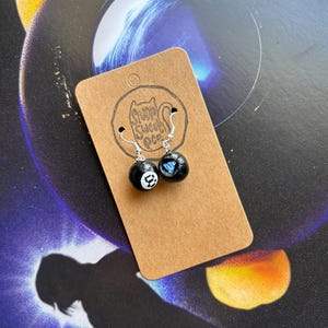 May include: A pair of black and white 8-ball earrings with silver hooks. One earring has a white number 8, the other a blue heart. They are displayed on a brown card with a logo reading "Stump Sweet Pea".