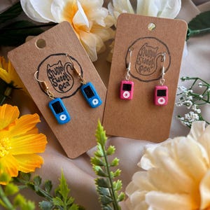 May include: A pair of dangle earrings featuring miniature iPods. One pair is blue and the other is pink. The earrings are displayed on a brown card with the text "Simply Sweet Pea".