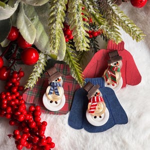May include: Three festive snowman decorations. Each snowman is crafted from a large white button body, a smaller button head, and a brown hat. They are adorned with scarves and set against red, blue, and plaid patterned backgrounds, perfect for Christmas.