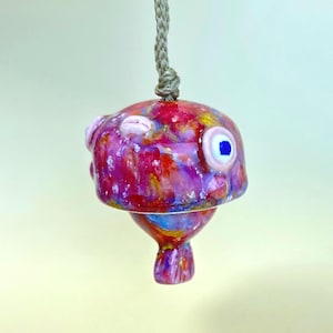 May include: A ceramic pufferfish ornament featuring a glossy, multi-colored glaze in shades of pink, red, purple, and blue. The fish has a prominent round eye with a blue pupil and is suspended from a thick, light brown braided rope cord.