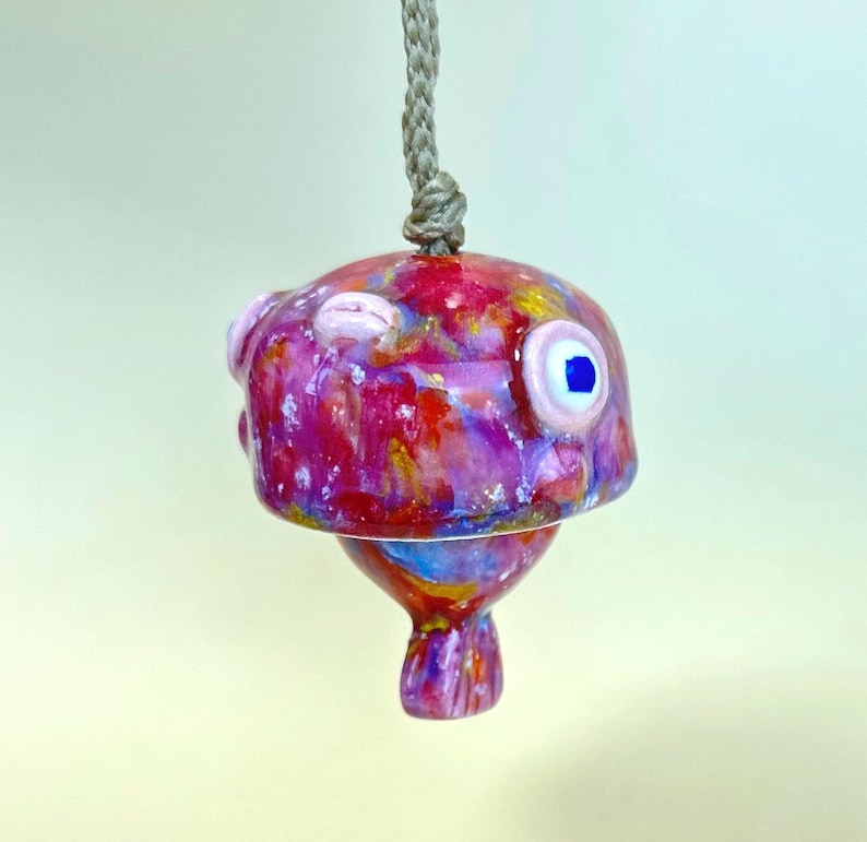 May include: A ceramic pufferfish ornament featuring a glossy, multi-colored glaze in shades of pink, red, purple, and blue. The fish has a prominent round eye with a blue pupil and is suspended from a thick, light brown braided rope cord.