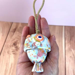 May include: A ceramic fish-shaped ornament featuring a glossy glaze with a mottled pattern of light blue, orange, and yellow spots. The ornament has a prominent orange eye with a dark blue center and is suspended by a thick, braided tan rope loop.