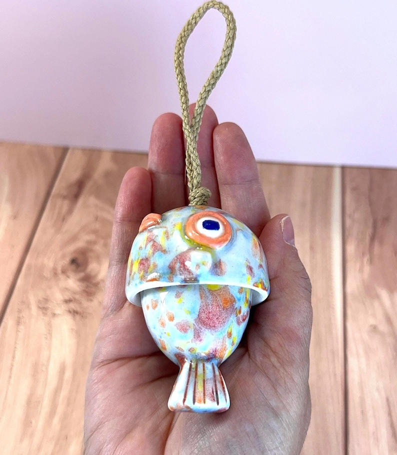 May include: A ceramic fish-shaped ornament featuring a glossy glaze with a mottled pattern of light blue, orange, and yellow spots. The ornament has a prominent orange eye with a dark blue center and is suspended by a thick, braided tan rope loop.