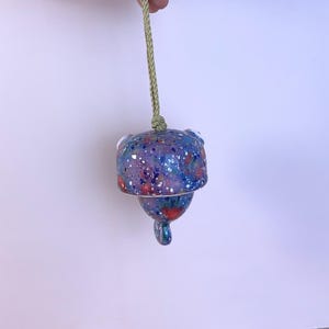 May include: A hanging ceramic bell featuring a speckled finish in shades of blue, purple, and red with white flecks. The bell is suspended by a light green braided cord attached to the top, and it has a small, rounded clapper extending from the bottom.