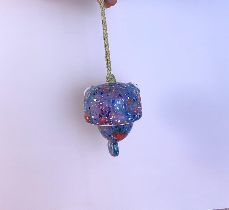 May include: A hanging ceramic bell featuring a speckled finish in shades of blue, purple, and red with white flecks. The bell is suspended by a light green braided cord attached to the top, and it has a small, rounded clapper extending from the bottom.