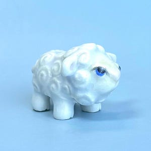 Könnte beinhalten: Eine kleine Schaf-Figur aus weißer Keramik mit einem texturierten, lockigen Wollmuster auf Körper und Kopf. Das Schaf hat eine glatte, glänzende Oberfläche und ist mit einem einzelnen, deutlich sichtbaren leuchtend blauen Auge auf vier kurzen Beinen versehen.