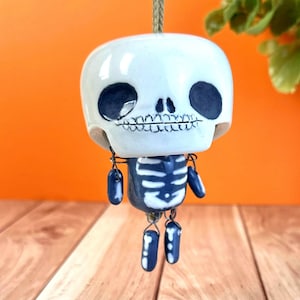 May include: A white ceramic skeleton ornament with a blue spine and limbs. The skull has large black eye sockets and a detailed jawline. The ornament is suspended by a green string, perfect for Halloween or Day of the Dead decor.