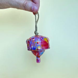 May include: A ceramic bell ornament featuring a vibrant, multi-colored glaze with swirling patterns of red, blue, yellow, and purple. The bell is suspended by a thick, natural-colored twisted rope cord attached to the top of the rounded ceramic body.