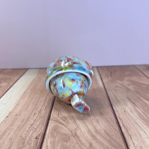 May include: A small, rounded ceramic object with a glossy finish, featuring a mottled pattern of light blue, yellow, and reddish-brown speckles. The piece has a distinct, curved protrusion extending from the base and a smooth, rounded top section.