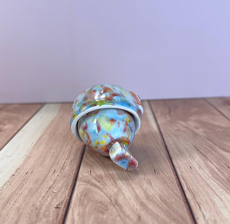 May include: A small, rounded ceramic object with a glossy finish, featuring a mottled pattern of light blue, yellow, and reddish-brown speckles. The piece has a distinct, curved protrusion extending from the base and a smooth, rounded top section.