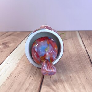 May include: A bell-shaped ornament featuring a multi-colored glass bird clapper inside. The exterior of the bell is a glossy, mottled red and white finish. A small loop of gold-colored cord is attached to the top for hanging purposes.