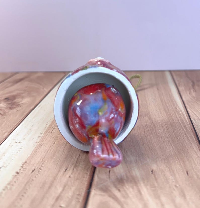 May include: A bell-shaped ornament featuring a multi-colored glass bird clapper inside. The exterior of the bell is a glossy, mottled red and white finish. A small loop of gold-colored cord is attached to the top for hanging purposes.