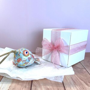 May include: A ceramic fish ornament with a mottled blue, orange, and yellow glaze, featuring a prominent eye and a braided cord, positioned next to a white square gift box wrapped with a translucent pink ribbon tied in a bow.