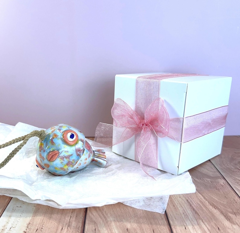 May include: A ceramic fish ornament with a mottled blue, orange, and yellow glaze, featuring a prominent eye and a braided cord, positioned next to a white square gift box wrapped with a translucent pink ribbon tied in a bow.