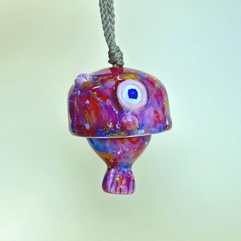 May include: A hanging ceramic ornament shaped like a fish, featuring a vibrant, multi-colored glaze in shades of magenta, red, blue, and gold. The fish has a single prominent eye with a blue pupil and is suspended by a thick, braided gray cord.