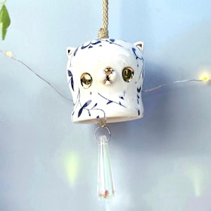 May include: A white ceramic cat-shaped wind chime with blue floral patterns and gold accents. The cat has large, round eyes and a small nose. A clear, teardrop-shaped crystal hangs below, suspended by a silver ring and a beige cord.