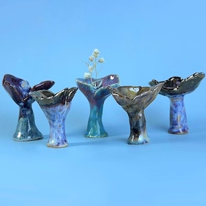 May include: Five small, hand-sculpted ceramic vessels with flared, organic rims and narrow bases. Each piece features a unique glaze finish in shades of deep purple, blue, brown, and teal. One vessel contains a small, delicate sprig of dried white flowers.