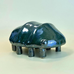May include: A ceramic sculpture shaped like a beetle with a glossy, dark teal and blue iridescent glaze. The object features a segmented, rounded body with a smooth surface and stands on six short, sturdy legs, reflecting light across its curved form.
